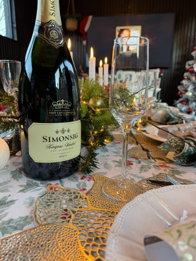 Feestelijk gedekte kersttafel met een fles Kaapse MCC, een leeg champagneglas, kaarslicht en kerstversiering